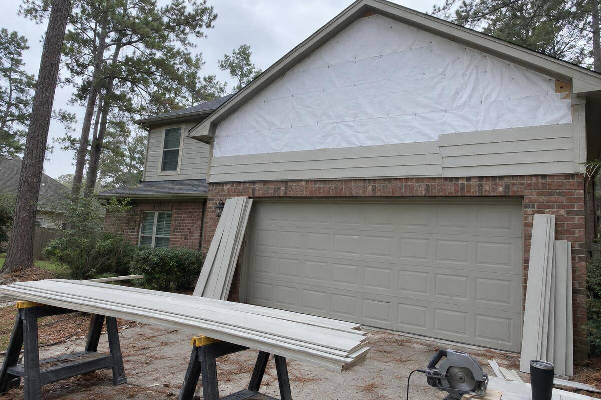 An upper gable stripped to house wrap, with primed lap planks stacked on sawhorses in the driveway