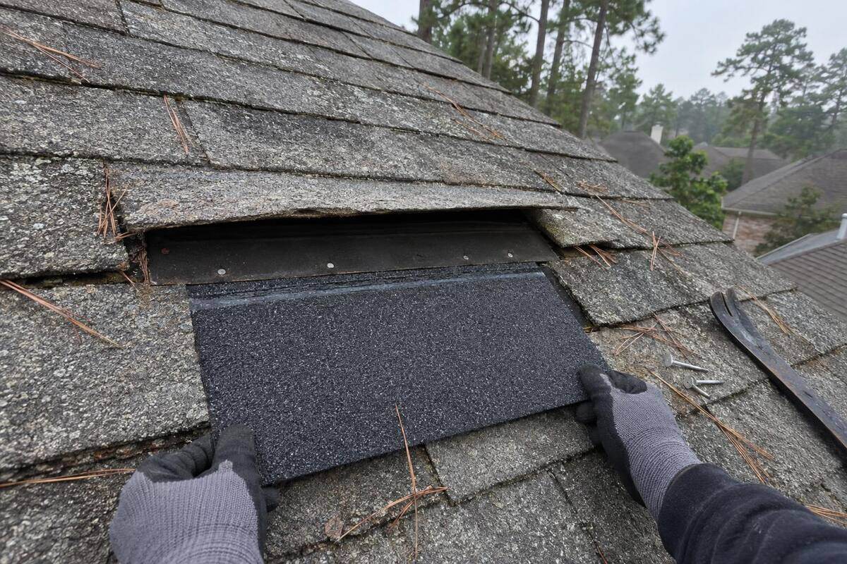Gloved hands sliding a darker replacement shingle under a lifted tab on a weathered roof